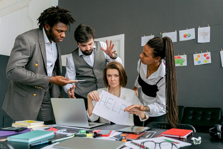 Office scene with diverse colleagues around a stressed woman holding a sign that says 'HELP'. Focus on teamwork dynamics.