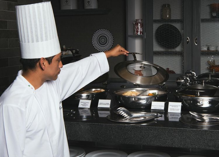 Chef in white uniform opening lid of buffet dish in modern kitchen setting.