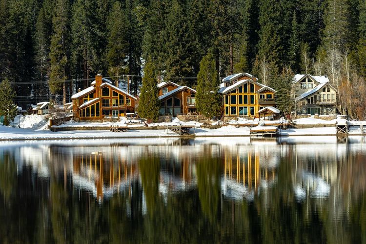 Charming wooden cabins reflect on a tranquil snow-covered lakeside, surrounded by tall pine trees.