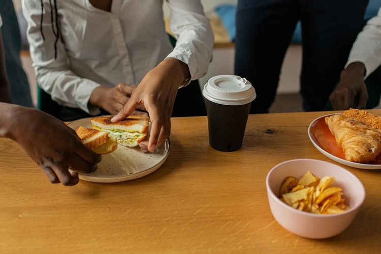 Close-up of a diverse group sharing sandwiches, coffee, and snacks at a wooden table.